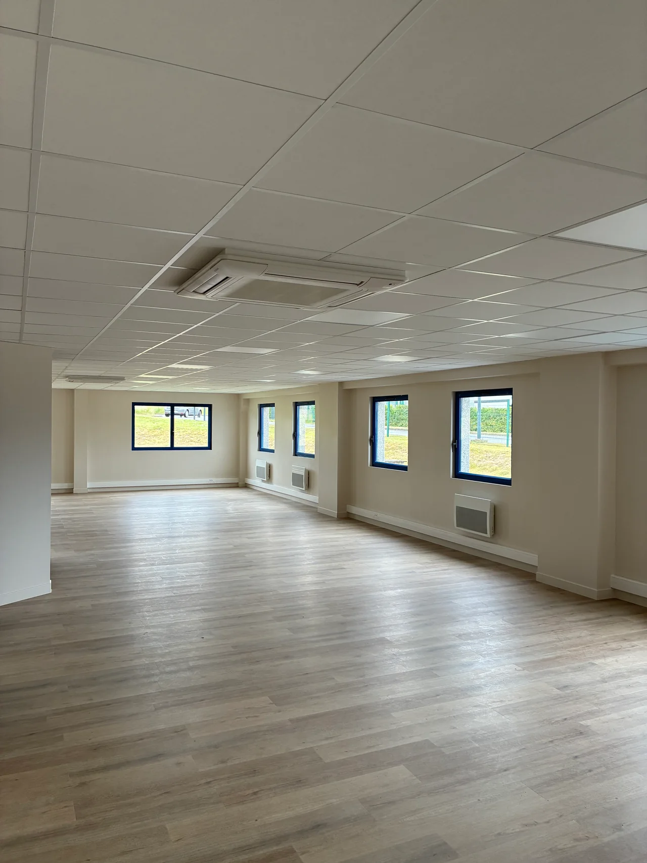 Grand espace de bureaux avec sol aspect bois, faux plafond et fenêtres sur l’extérieur.